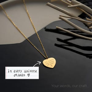 May include: A gold-colored necklace with a heart-shaped pendant. The pendant is engraved with the words "in every universe 09/05/23 ♡". The necklace is displayed on a black and gray surface with decorative wooden sticks.