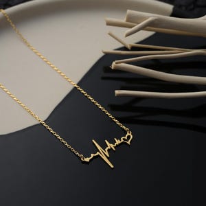 May include: A gold-colored necklace featuring a heartbeat design, with a chain and a pendant shaped like an electrocardiogram. The necklace is displayed on a black and white surface, with decorative wooden sticks in the background.
