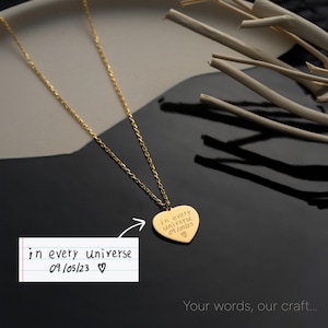 May include: A gold-colored necklace with a heart-shaped pendant. The pendant is engraved with the words "in every universe 09/05/23 ♡". The necklace is displayed on a black and gray surface with decorative wooden sticks.