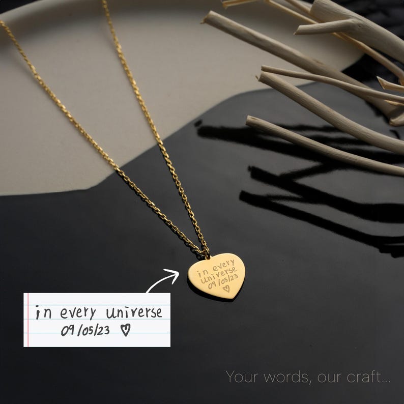 May include: A gold-colored necklace with a heart-shaped pendant. The pendant is engraved with the words "in every universe 09/05/23 ♡". The necklace is displayed on a black and gray surface with decorative wooden sticks.