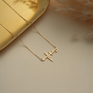 May include: A delicate gold-colored necklace featuring a heartbeat design with a small heart at the end. The necklace is displayed on a gold-colored tray, with a soft, neutral background.