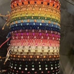 May include: A collection of macrame bracelets in a rainbow of colours, including red, orange, yellow, green, blue, and purple. Each bracelet is intricately woven with small beads, creating a textured design. The bracelets are stacked together.