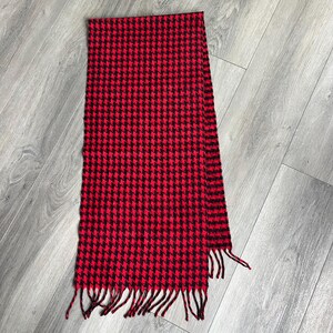 May include: A rectangular scarf featuring a repeating houndstooth pattern in vibrant red and black. The scarf is finished with short, matching red and black fringe along the bottom edge, providing a textured look to the overall design.