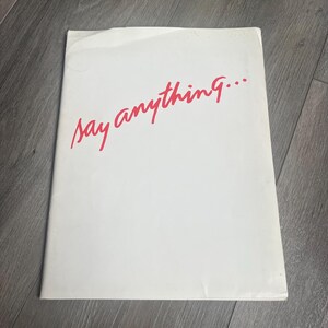 May include: A rectangular white paper item featuring the text say anything... written in a cursive red font. The surface of the paper appears slightly creased near the top edge, and the overall orientation is vertical with a clean white background.