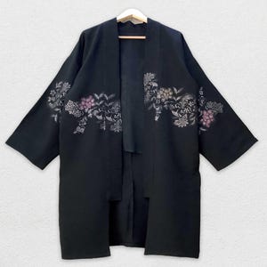 May include: A black kimono-style robe with floral embroidery in shades of pink, silver, and gold. The robe has a loose, open front design and three-quarter length sleeves. The floral pattern is concentrated across the chest and on the sleeves.