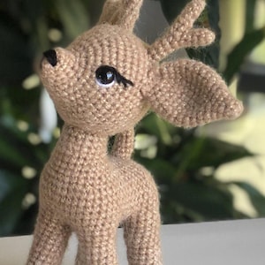 May include: A crocheted fawn with brown fur and large, black eyes. The fawn has antlers and is standing on a white surface.