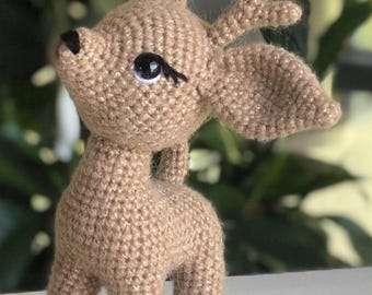 May include: A crocheted fawn with brown fur and large, black eyes. The fawn has antlers and is standing on a white surface.