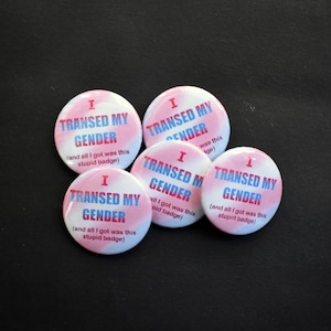 May include: A collection of five round buttons with the text "I TRANSED MY GENDER" in a pink and blue gradient design. The text "(and all I got was this stupid badge)" is also included on the buttons. The buttons are on a black surface.