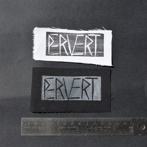 Puede incluir: Dos parches de tela en blanco y negro con la palabra "PERVERT" impresa en blanco sobre fondo negro y negro sobre fondo blanco.