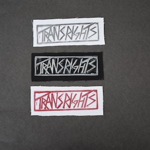 Könnte beinhalten: Drei Stoff-Patches mit dem Aufdruck "Trans Rights" in Weiß, Silber und Rot auf schwarzem, weißem und weißem Hintergrund.