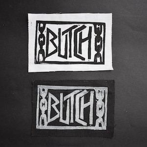 Può includere: Due toppe rettangolari con la parola "BUTCH" in grassetto e in stile. Una è bianca con inchiostro nero, l'altra nera con inchiostro argento. La parola è incorniciata da un bordo decorativo.