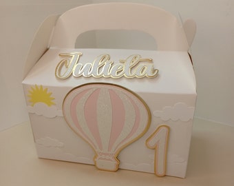 May include: A white favour box with a handle, decorated with a pink and white hot air balloon, a yellow sun, white clouds, and gold lettering that says "Julieta 1".