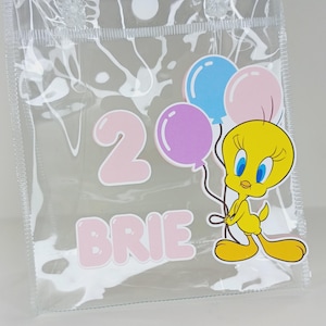 Puede incluir: Bolsa de mano de plástico transparente con asa, diseñada para un segundo cumpleaños. El diseño incluye el número "2", el nombre "BRIE" y un personaje de dibujos animados Piolín sosteniendo globos azules, rosas y morados.