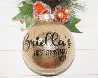 以下が含まれることがあります： 「Briella's First Christmas 2023」の黒い文字が入った、金色のグリッタークリスマスオーナメント。オーナメントは金の紐で吊り下げられており、お祝いの緑とベリーに囲まれています。