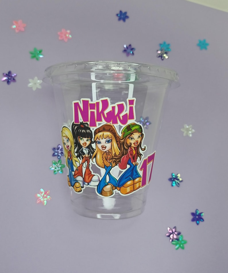 May include: Clear plastic cup with a pink and purple sticker featuring a group of cartoon girls with the name "Nikki" printed on the sticker.