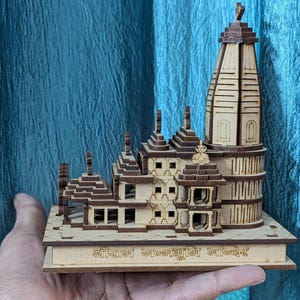 May include: A detailed wooden model of a temple, with intricate architectural features. The model is mainly light brown with darker brown accents, showing multiple levels, towers, and decorative elements. The base has text in a foreign language.