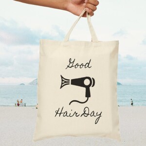 May include: A white canvas tote bag with black text and a hairdryer graphic. The text reads "Good Hair Day".