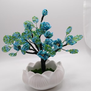 May include: A decorative tree sculpture featuring branches made of twisted dark wire adorned with small blue and green glass beads. The tree is set in a white ceramic pot shaped like an open lotus flower, with green material visible at the base.