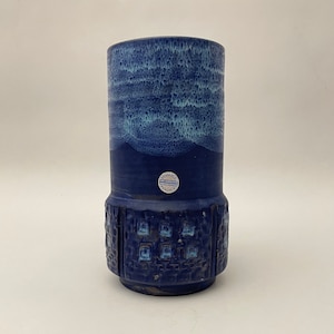 May include: A blue ceramic vase with a cylindrical shape. The top has a textured, watercolour-like effect, fading from light to dark blue. The lower part features a geometric pattern. A small sticker is visible near the base.