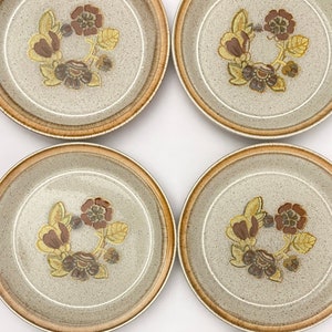 May include: Six ceramic plates with a speckled, light gray surface. Each plate features a brown and yellow floral design in the center, surrounded by a brown border. The plates are arranged in a grid pattern, showcasing a vintage tableware set.