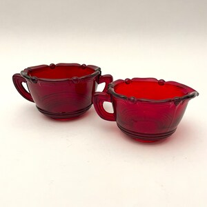 May include: Two ruby red glass teacups with handles. The cups have a scalloped rim with small beaded accents and a ribbed design on the lower portion. The cups are set against a white background.