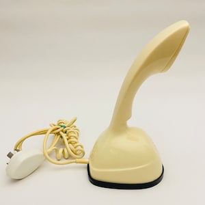 Könnte beinhalten: Cremefarbenes Vintage-Telefon mit gebogenem Hörer und Sockel. Das Telefon hat einen schwarzen Sockel und ein Spiralkabel. Ein weißer Stecker ist am Kabel befestigt. Das Telefon ist ein Retro-Design.