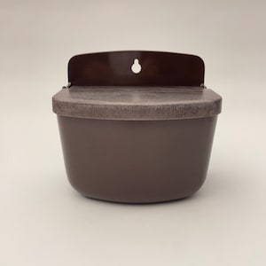 May include: A brown, wall-mounted container with a lid and a small, curved backplate. The container has a rounded shape and a textured surface on the lid. A hole is present on the backplate for hanging. The container is made of plastic.