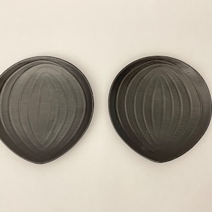 Könnte beinhalten: Zwei dunkelgraue, oval geformte Teller mit einem strukturierten, geriffelten Design. Die Teller haben eine leicht unregelmäßige Form und werden vor einem neutralen Hintergrund präsentiert. Die Teller sind wahrscheinlich zum Servieren von Speisen.