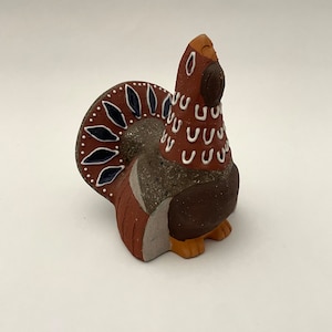 May include: A ceramic bird figurine with a brown body, a speckled gray chest, and a tail with dark blue and white accents. The bird has a brown head with white markings and is posed upright.