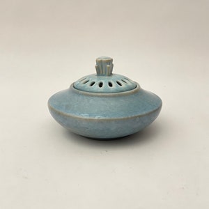 Może przedstawiać: Jasnoniebieski ceramiczny palnik do kadzideł z pokrywką z wieloma małymi otworami i ozdobnym uchwytem. Palnik ma zaokrągloną podstawę i pokrywkę, która dobrze pasuje. Prosty i elegancki design.