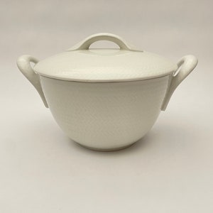 以下が含まれることがあります： 蓋付きの白いセラミック製サービングボウル。ボウルには2つの湾曲したハンドルと、蓋にループハンドルが付いています。ボウルと蓋の表面には、微妙なテクスチャパターンがあります。ボウルは食べ物を提供するように設計されています。