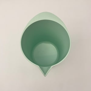 May include: A light green plastic measuring cup with a spout. The cup has a wide, open top and a rounded shape, with a pointed spout for easy pouring. The interior of the cup is the same color as the exterior.