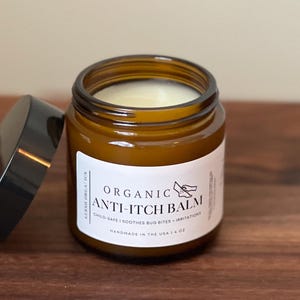 May include: A brown glass jar with a black lid, containing a white balm. The jar has a white label with the words "Organic Anti-Itch Balm" and a small bird illustration. The jar is on a wooden surface.