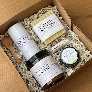 May include: A gift box containing organic personal care items. The box includes a deodorant, shampoo bar, toothpaste, lip balm, and tallow balm. The products are in a cardboard box with paper filler, and a small wooden spoon.
