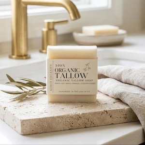 May include: A bar of organic tallow soap with a label stating "100% ORGANIC TALLOW". The soap sits on a textured stone surface, with a sprig of leaves and a folded linen towel. A gold tap is visible in the background.