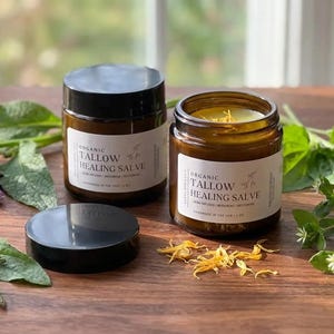May include: Two amber glass jars with black lids containing organic tallow healing salve. One jar is closed, and the other is open, revealing the yellow salve inside. The white labels feature the text Organic Tallow Healing Salve.