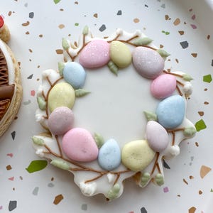 May include: A white sugar cookie decorated with a wreath of pastel colored eggs and brown branches. The cookie is on a white background with brown and gray speckles.