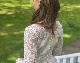 Könnte beinhalten: Ein weißes Spitzen-Hochzeitskleid mit langen Ärmeln und einem Knopfleistenrücken. Das Kleid hat ein tailliertes Oberteil und einen vollen Rock. Die Braut trägt einen weißen Spitzenhut.