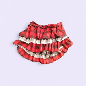 May include: A tiered skirt with a red and black plaid pattern, accented by layers of white and gray fabric. The skirt has a ruffled design and a gathered waist, creating a playful and stylish look. The skirt is made of a lightweight material.