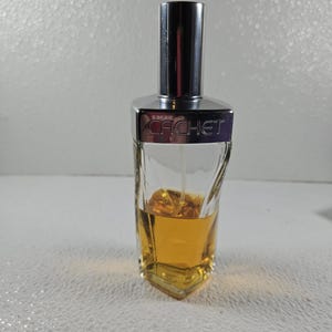 May include: A clear glass perfume bottle with a silver cap and the word "CACHET" printed on the front. The bottle contains a golden-colored liquid. The bottle has a unique, angular shape.
