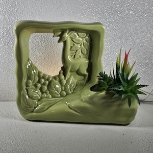 May include: A light green ceramic lamp featuring a relief sculpture of a deer standing among foliage. The structure includes an open rectangular frame and a built-in planter section on the right side, which contains several artificial succulent plants.
