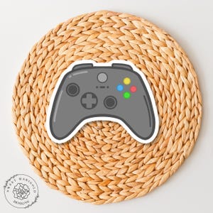 May include: A gray video game controller with black buttons and a white background. The controller has four colored buttons: green, blue, yellow, and red.