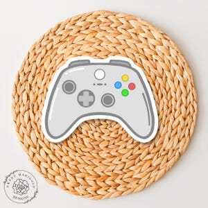 May include: A white and gray video game controller with colorful buttons on a woven straw background.