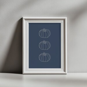 May include: A framed wall art piece featuring a dark blue background with three vertically aligned white line art illustrations of pumpkins. The artwork is enclosed in a simple white frame with a clean, rectangular border design.