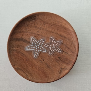 May include: Two clear starfish stickers with white dotted details are displayed on a round, wooden plate. The plate has a rich, brown color and a natural wood grain. The stickers are a nautical-themed craft supply.
