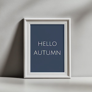 Puede incluir: Un marco de fotos rectangular blanco que contiene una impresión azul oscuro con el texto blanco HELLO AUTUMN centrado en el medio. El marco presenta un diseño de borde simple y limpio y se muestra contra una pared gris claro.