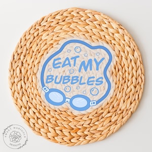 May include: A light brown woven placemat with a blue and white sticker that says "Eat My Bubbles" with a pair of blue goggles.