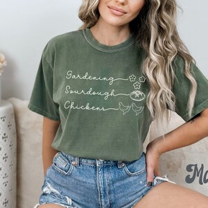 May include: A green t-shirt with the text "Gardening, Sourdough, Chickens" in white lettering. The shirt has a distressed look and is being worn by a person with long blonde hair.