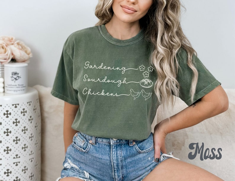 May include: A green t-shirt with the text "Gardening, Sourdough, Chickens" in white lettering. The shirt has a distressed look and is being worn by a person with long blonde hair.