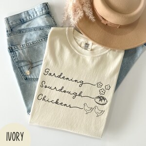 May include: Ivory t-shirt with the text "Gardening, Sourdough, Chickens" in black script. The shirt features a small illustration of a loaf of bread and two chickens.